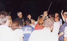 屋外で開催されたウジジ浜公園の交流会で、夜間複数の人々が円形に集まり、中央で棒状の物を持つ人物を囲んで談話やリジー・C・トゥループ号の釘に見入る乗組員の遺族の様子が写された写真