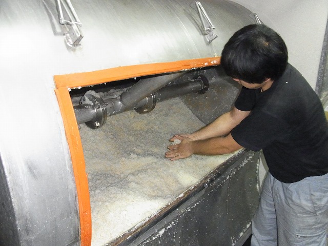 男性が白色の巨大な機械の開口部から腕を入れ、内部に広がる白い発酵中の麹を両手で触って状態を確認している、工場での作業風景写真