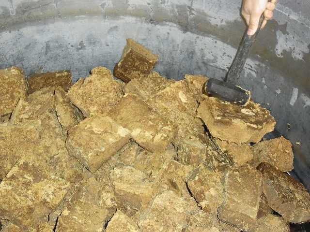 大きな容器の中に、茶色の角張ったブロック状の山積みになった黒糖をハンマーで割って割っている様子の写真