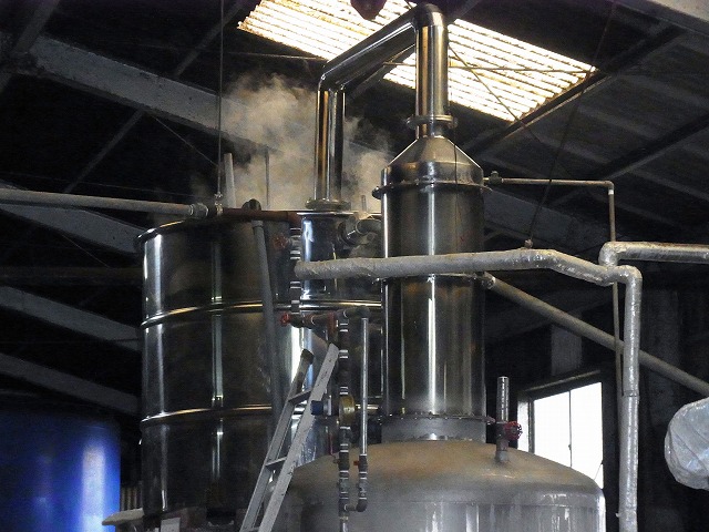 銀色の巨大な蒸留装置から水蒸気が上がっている製造工程の写真