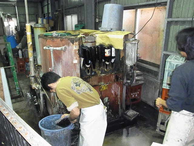 男性が、茶色の瓶が吊るされた機械の前にかがみ込み、茶色の瓶を青いバケツの中で洗浄する様子の写真