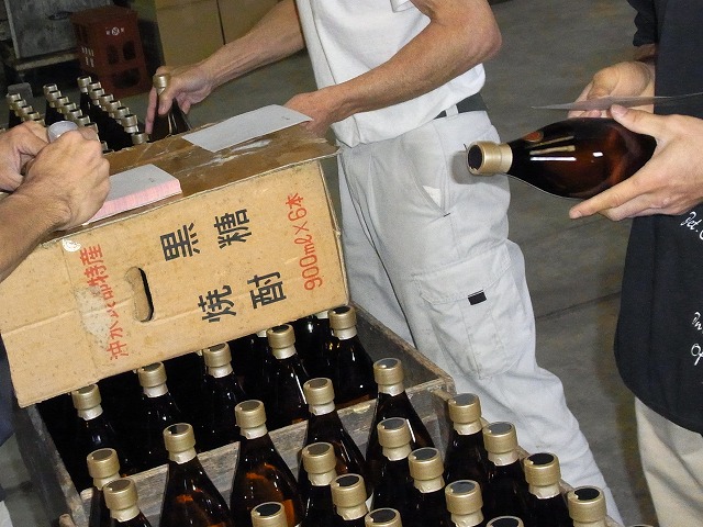 作業員3人が、ケースに入った茶色の瓶を囲み、金色のキャップがついた茶色の瓶にラベルを張っている酒造工場での作業写真