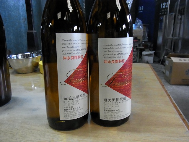 茶色い瓶に入った二本の濃いガラス瓶を、木製テーブルの上に立てて並べ、二本とも白地に赤いアクセントが入った「沖永良部特産・奄美黒糖焼酎」と書かれたラベルが貼られている製品のクローズアップ写真