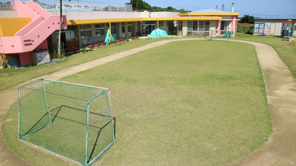 ピンク色の屋外階段が特徴的な平屋建ての保育園舎の前に広がる、芝生の園庭には小さなサッカーゴールが置かれ、芝生を囲むように子どもたちが走り回れるような土の小道が設けられている園庭の写真