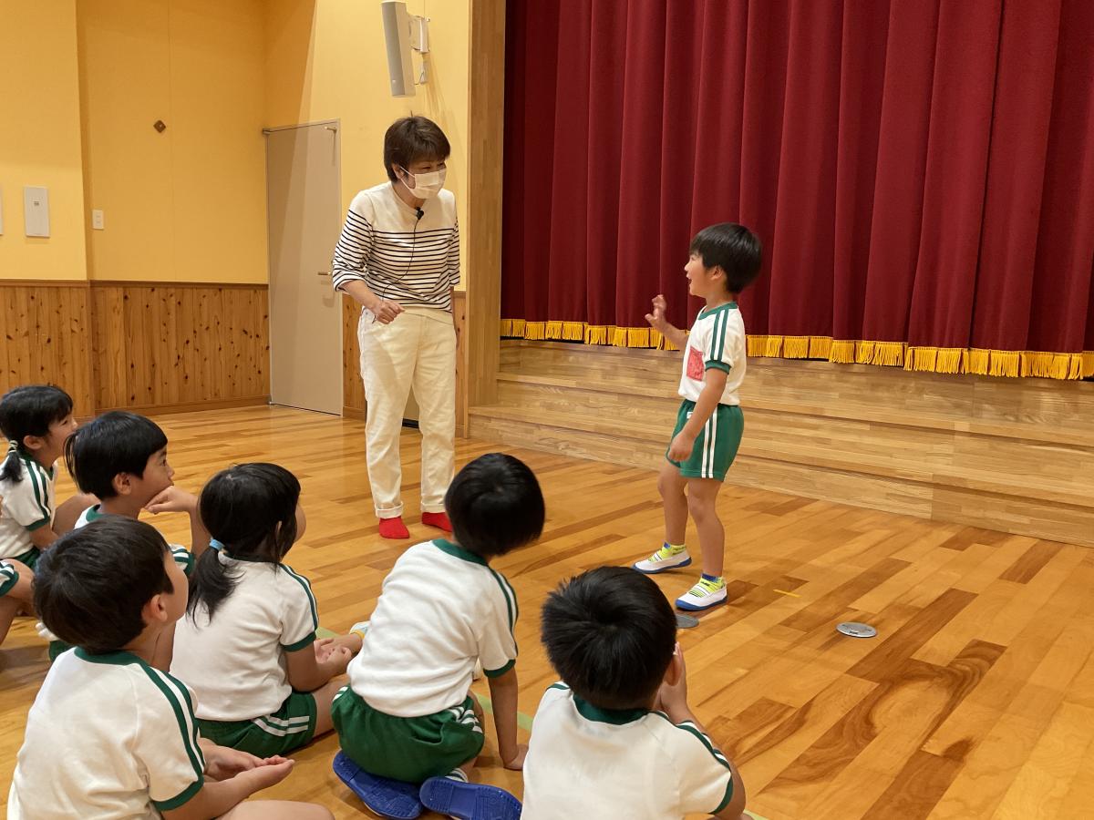 体操服姿の園児たちが床に座り、ステージ前に一人の男の子が手を挙げて先生に話しかけている様子の写真