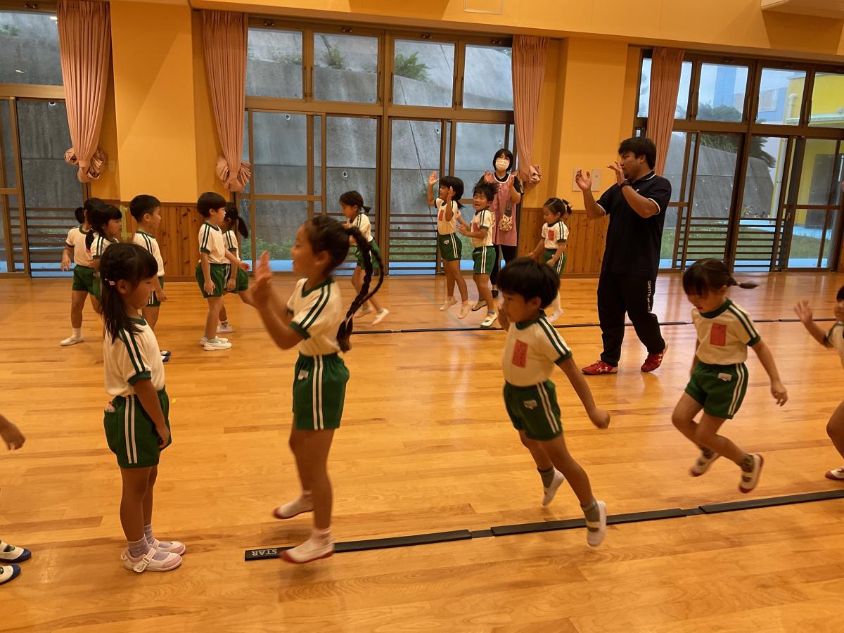 広い室内で、体操服を着た多くの園児が、黒いポロシャツの男性の先生の指導の下で活発に身体を動かしている様子の写真