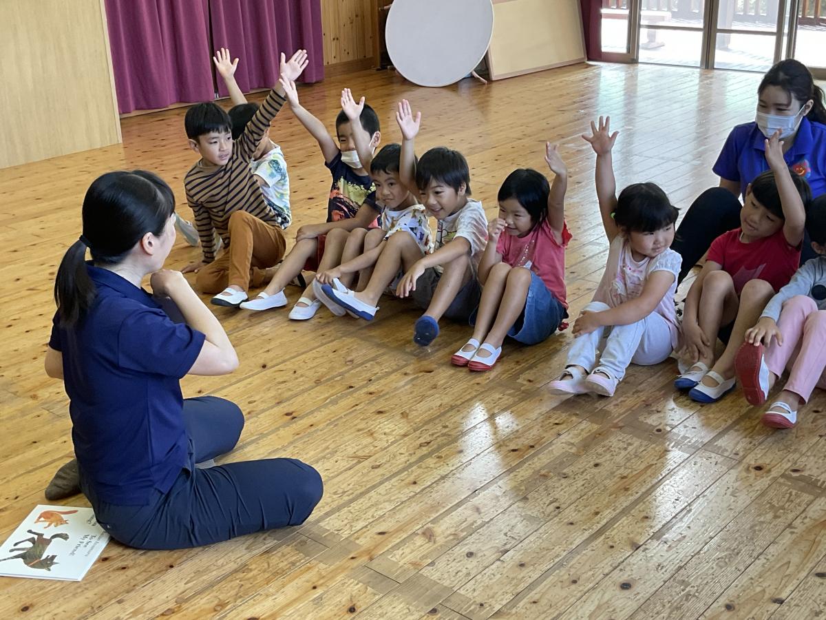 床の広間に座る紺色のポロシャツを着た先生の後ろに動物の絵本が置かれ、向かい合わせに座っている複数園児が手を挙げている写真
