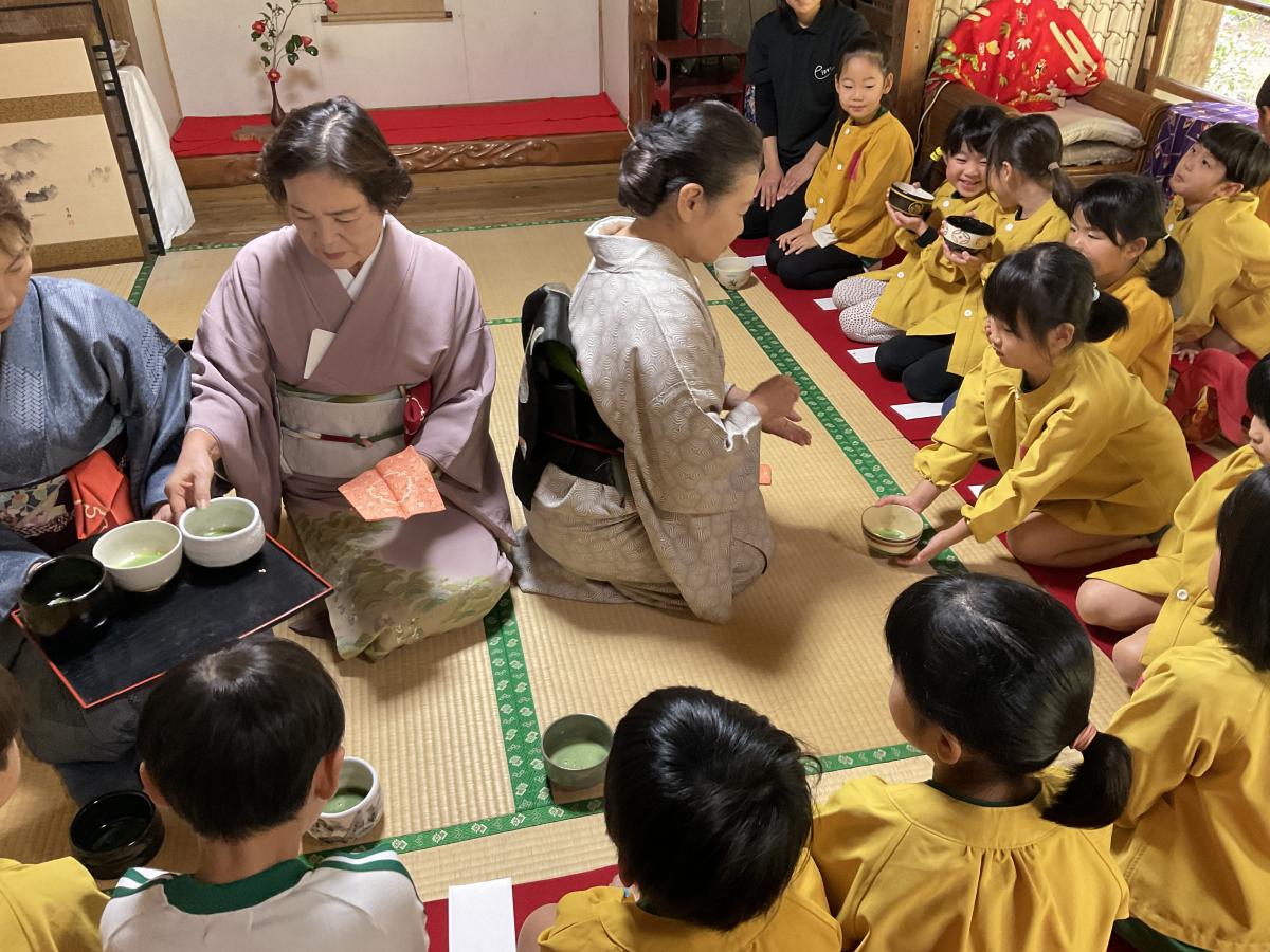 畳敷きの和室で、着物姿の3人の女性が前に座り、周りを黄色いスモックを着た園児たちが囲んで、お茶碗を手に持ったり受け取ったりしている茶道教室の写真
