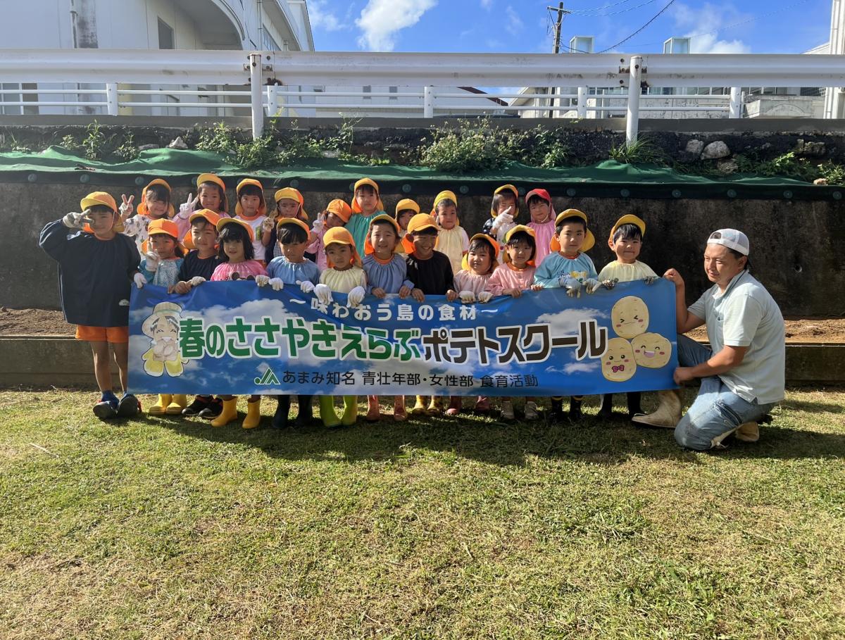 青空の下、広場のような場所で、黄色い帽子をかぶった20人以上の園児と横でしゃがんでいる男性一人が、「春のささやきえらぶポテトスクール」と書かれた青い横断幕を広げている記念写真