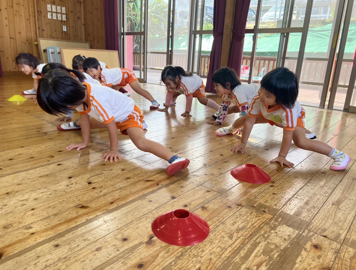 広々とした室内で、体操服を着た複数の園児が、両手を前についた低い体勢から大きく両足を広げているリズムトレーニングの写真