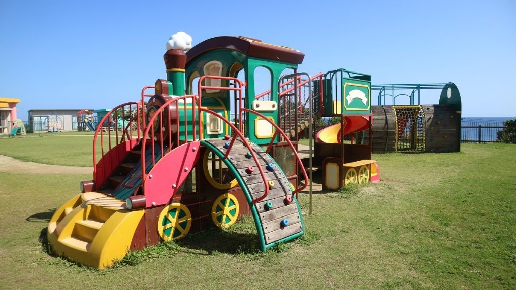 広々とした芝生の園庭の隅に、濃い緑色と赤、黄色の配色で、煙突から白い煙を出す蒸気機関車をモチーフにした、登り台や滑り台などが一体となった遊具の写真