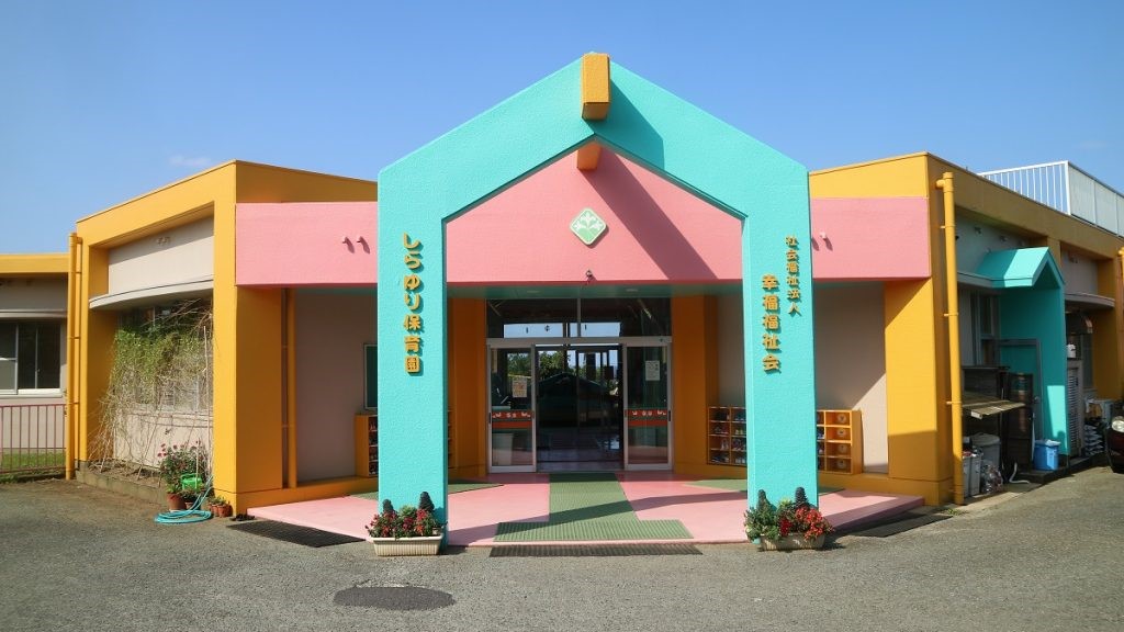 カラフルな建物が特徴的な薄いピンク色と黄色の平屋建ての建物にターコイズブルーの門があり、門柱には濃い黄色の文字で園名と「社会福祉法人 幸福福祉会」「しらゆり保育園」と書かれた外観写真