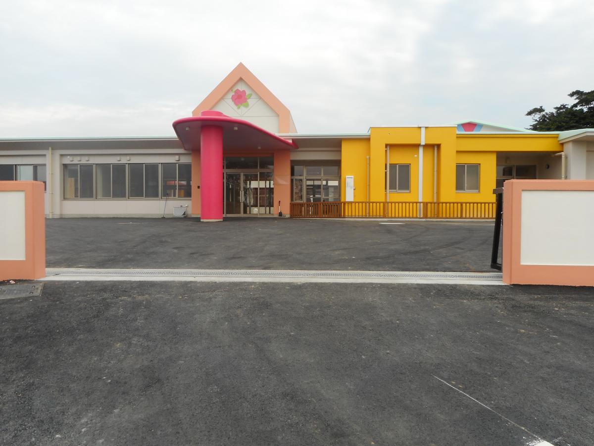 オレンジ色の縁取りの白い門柱の奥に建つ、中央の玄関ポーチが鮮やかなピンク色の大きな丸い柱と三角形の屋根で、右側の外壁が黄色の特徴的な平屋建ての認定こども園すまいるの外観写真