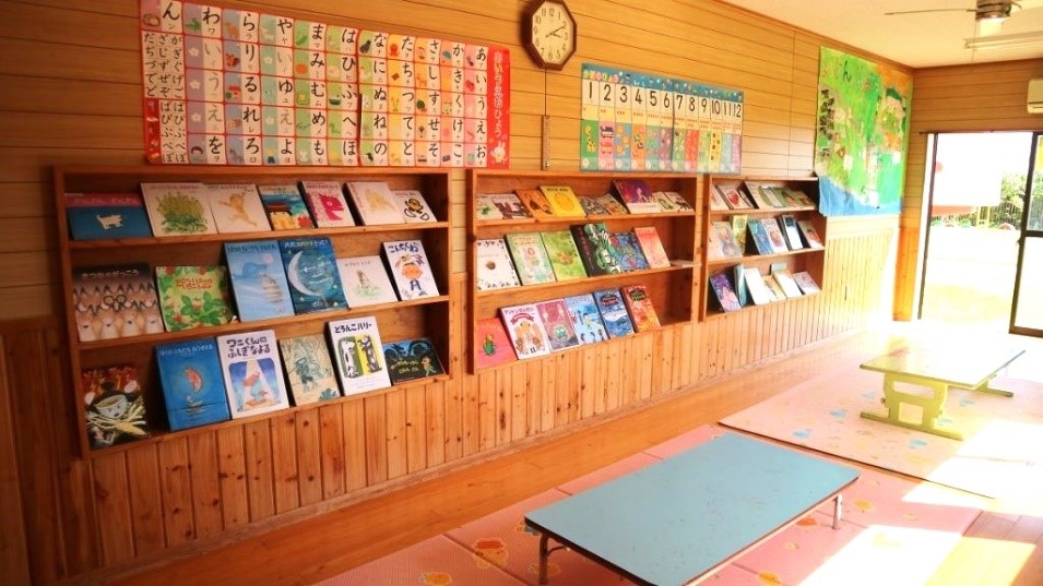 木目の壁と床の読書スペースに、ひらがなや数字のポスターの下に、様々な絵本が並べられた木製の棚があり、手前のピンク色のマットの上には低いテーブルが二つ置かれている図書室の写真